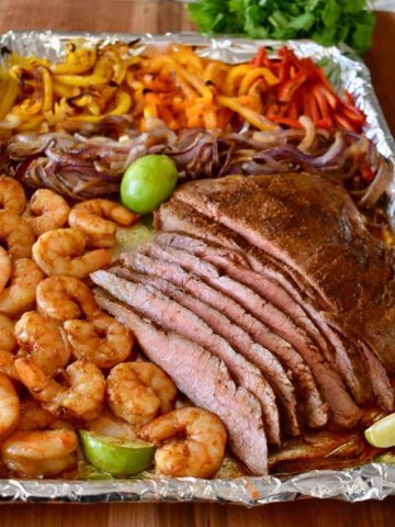 Sliced steak and shrimp fajitas on a lined baking sheet with bell peppers, onions, and lime.