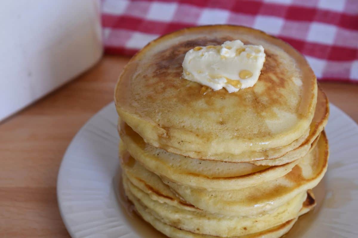 stack of pancakes on a white plate with butter and syrup and a checkered napkin in the background. 