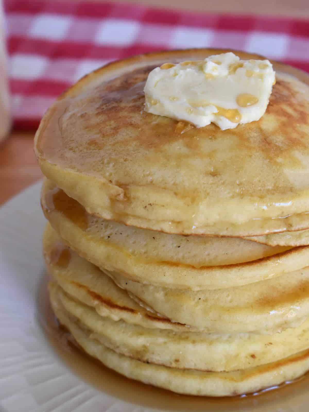 stack of pancakes on a white plate with butter and syrup on top and a checkered napkin in the background. 