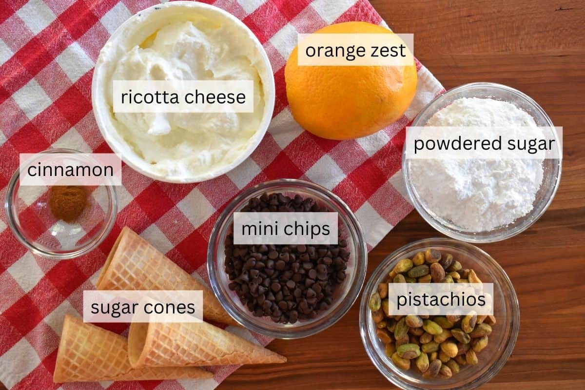 ingredients needed to make recipe including ricotta cheese, orange, powdered sugar, pistachios, chocolate chips, and sugar cones.