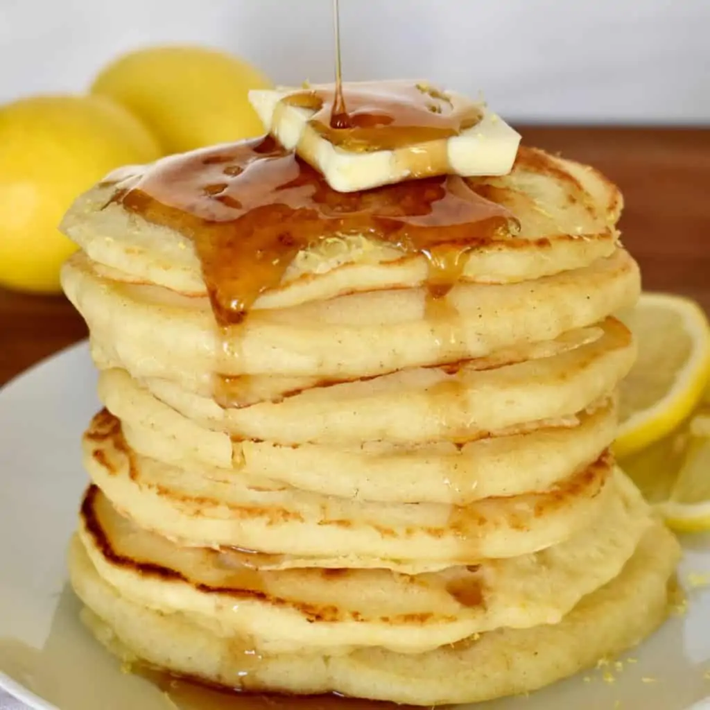 Lemon Ricotta Pancakes stacked on each other on a white plate with syrup being drizzle over top.