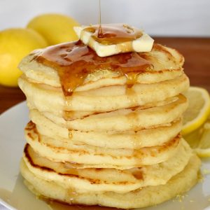 Lemon Ricotta Pancakes stacked on each other on a white plate with syrup being drizzle over top.
