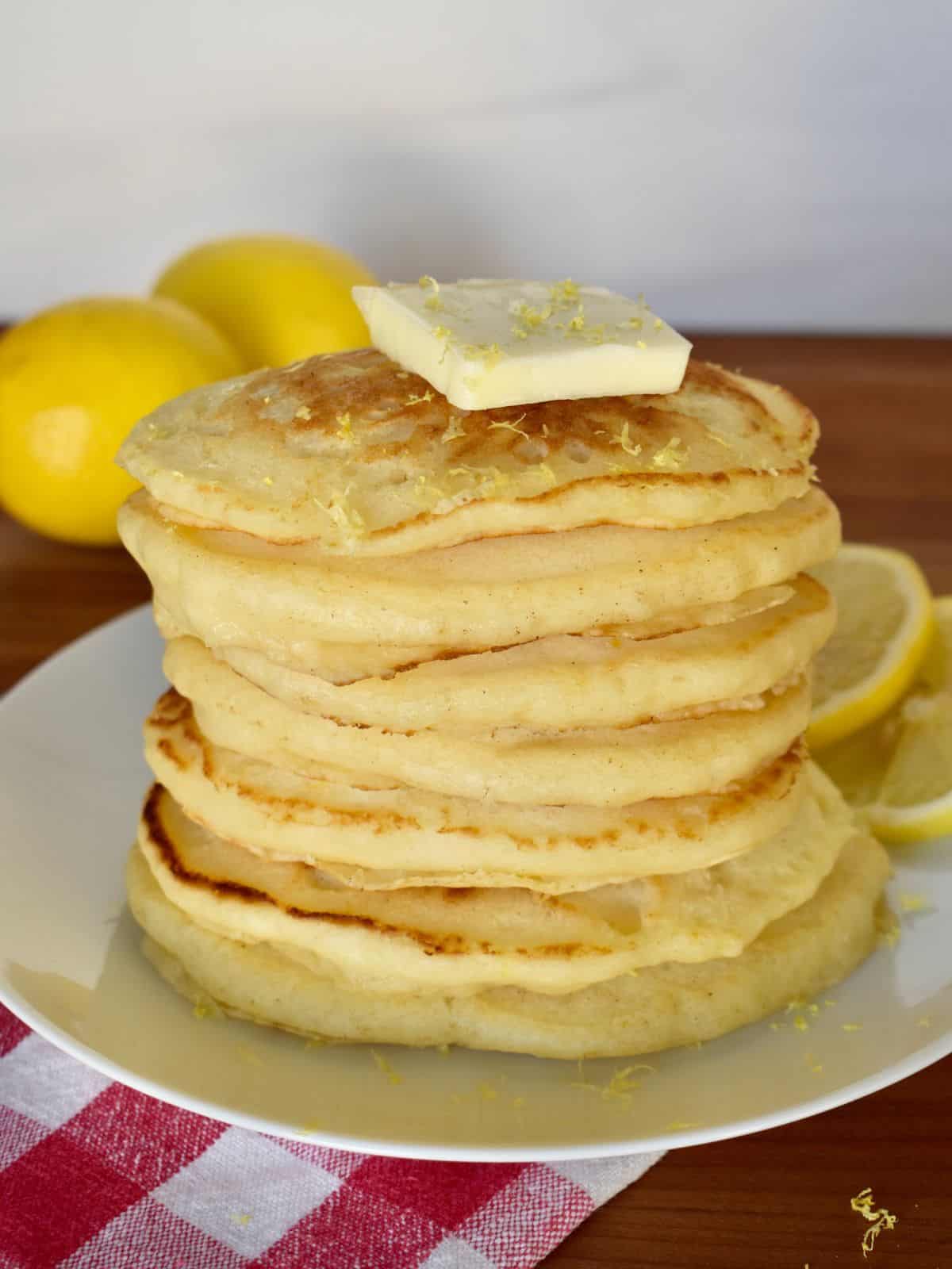 Stack of lemon ricotta pancakes with pancake mix on a white plate with butter on top and lemons in the background. 