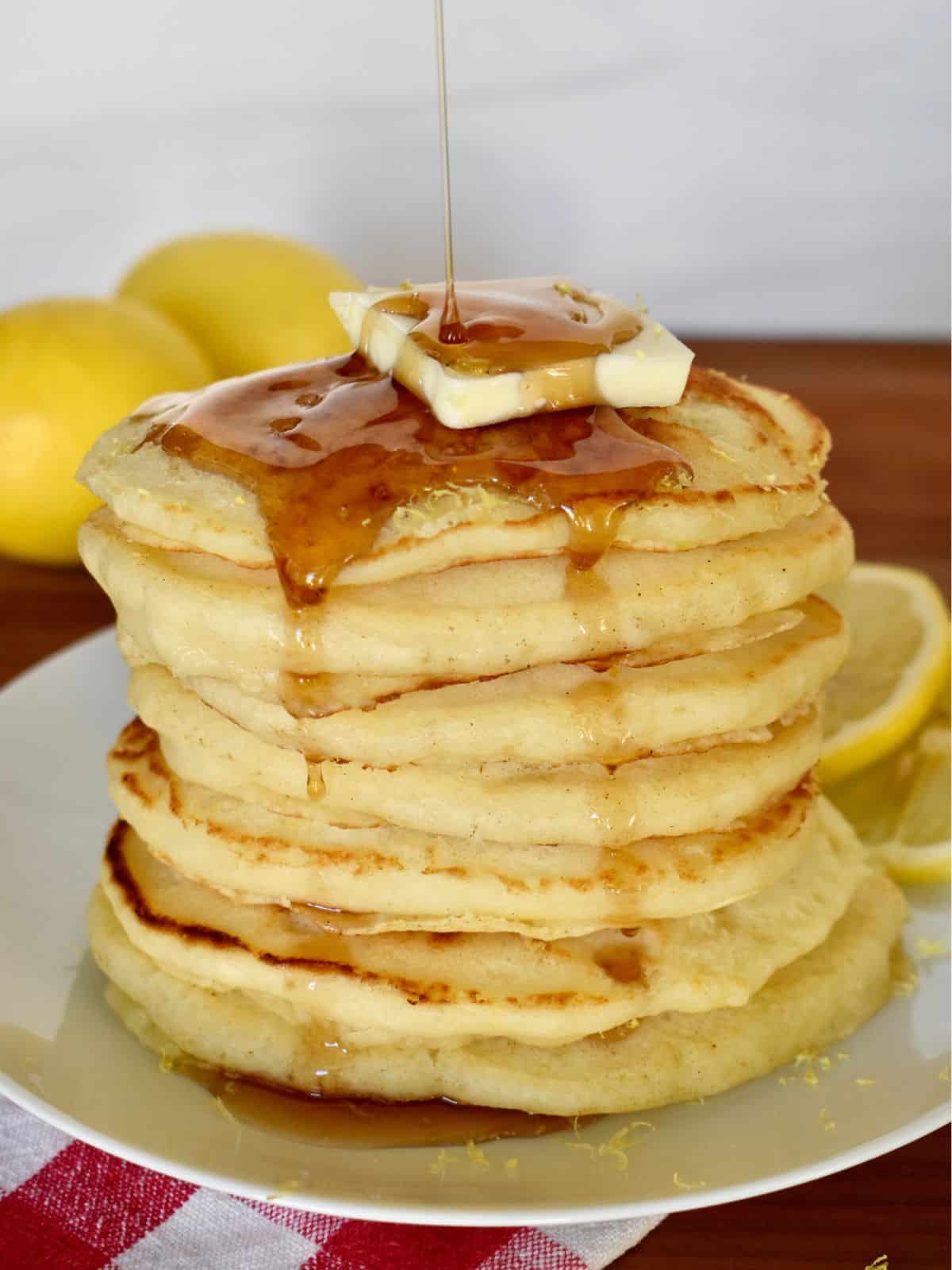 Stack of lemon ricotta pancakes on a white plate with butter on top and syrup drizzled over it. 