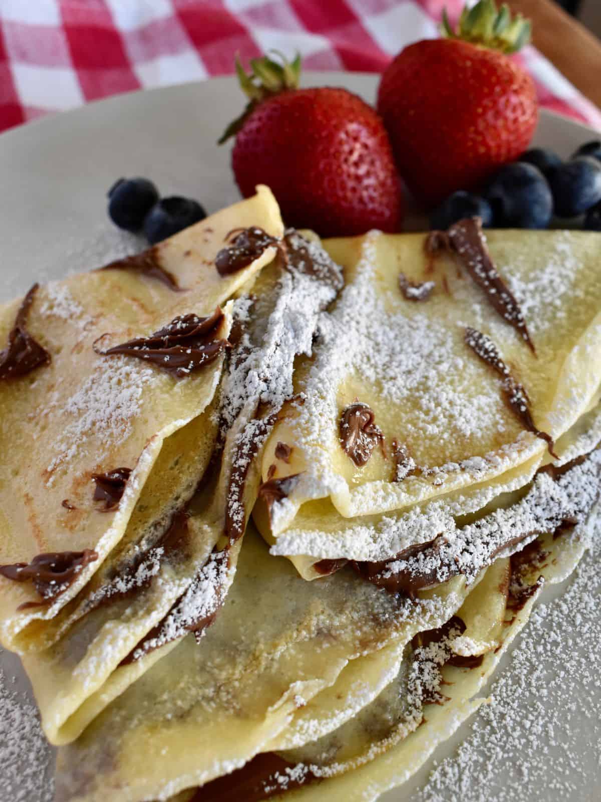 Nutella crepes on a white plate with powdered sugar on top and blueberries and strawberries. 