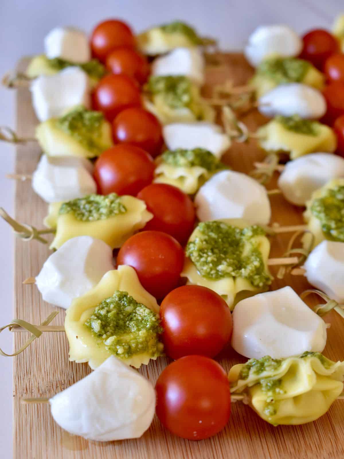 Angled view of easy pesto tortellini skewers made with tortellini, tomatoes, mozzarella, and pesto on a wood cutting board. 