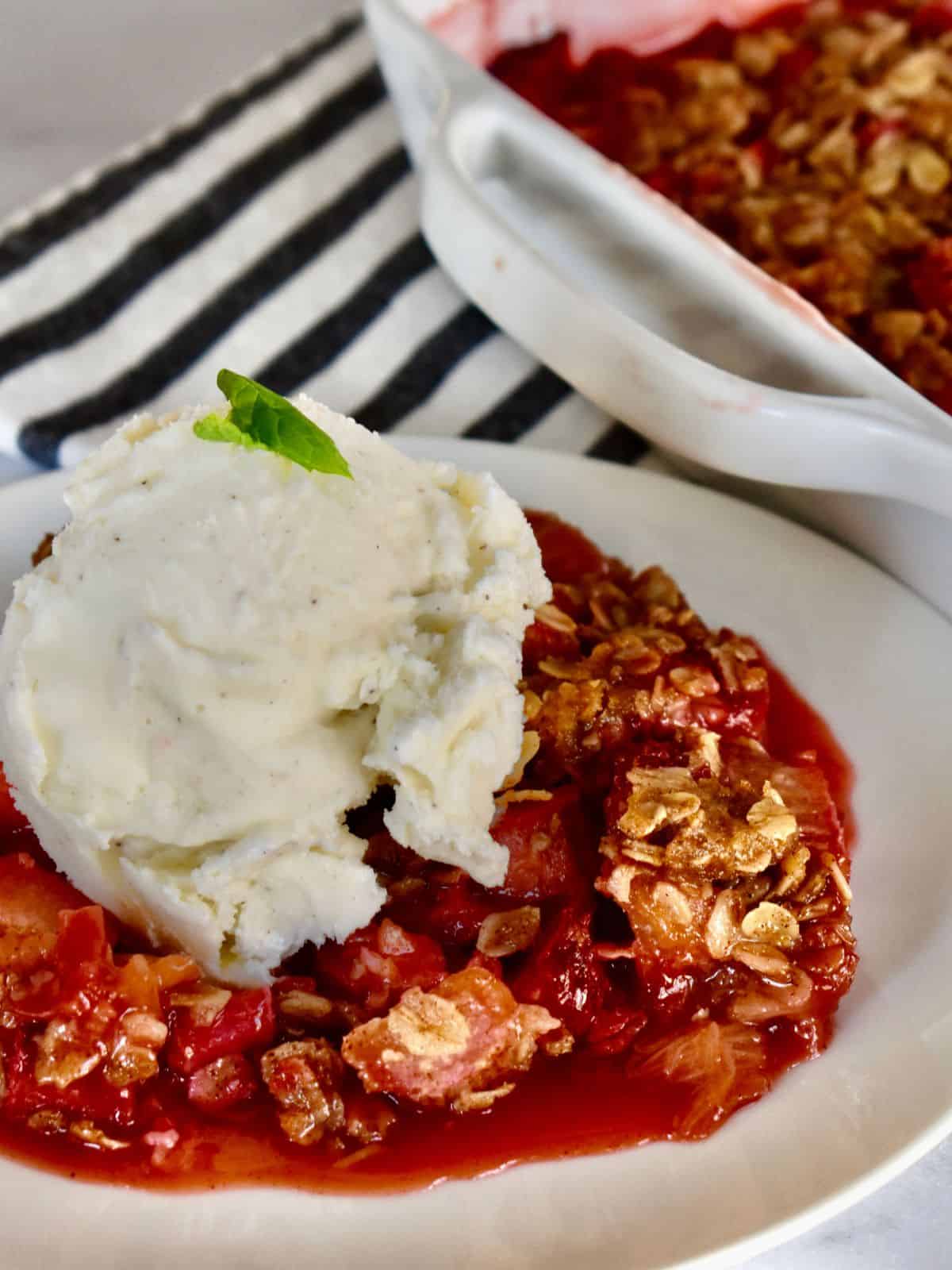 Strawberry rhubarb crisp in a white bowl with a scoop of vanilla ice cream on a striped napkin. 