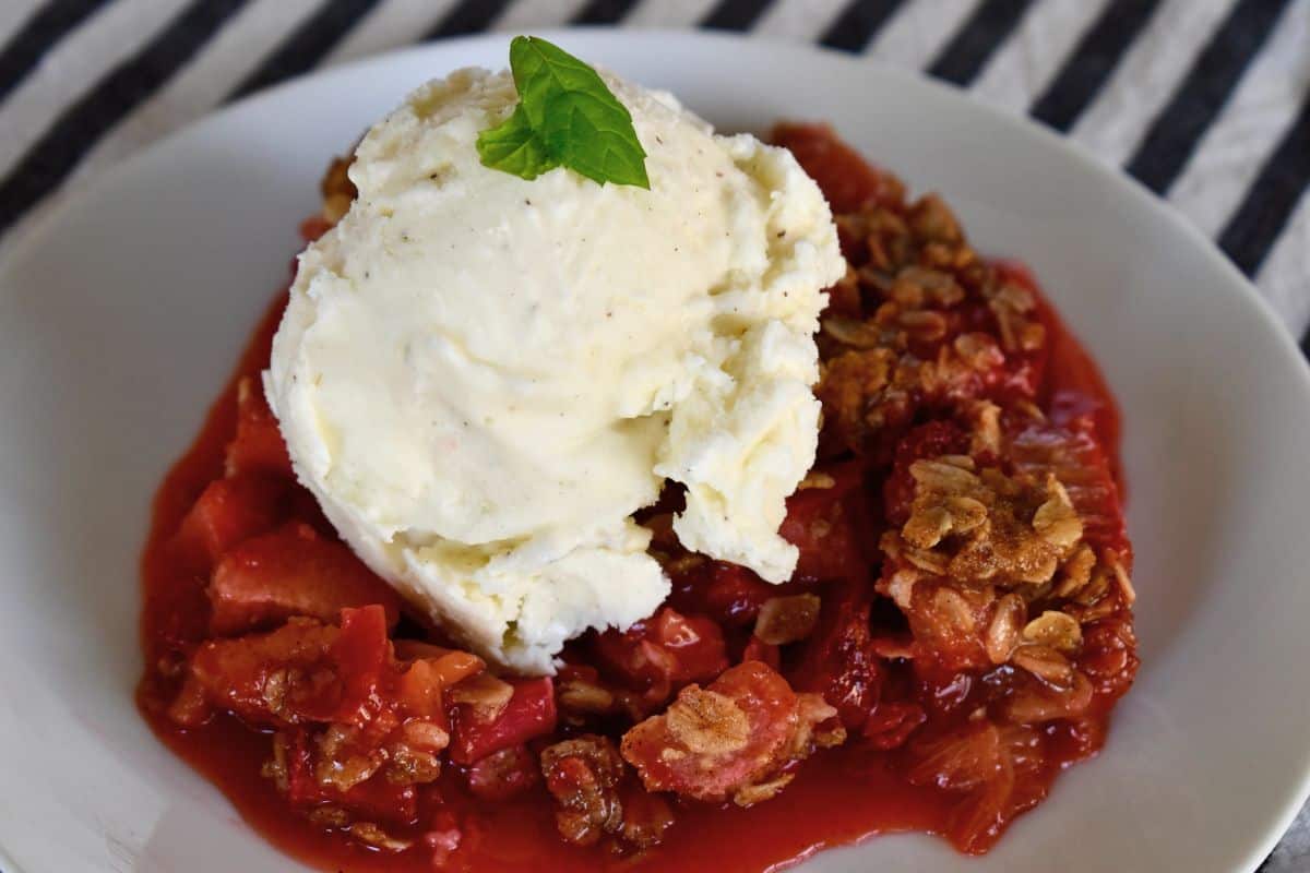 Strawberry rhubarb crisp in a white bowl with a scoop of vanilla ice cream on top. 