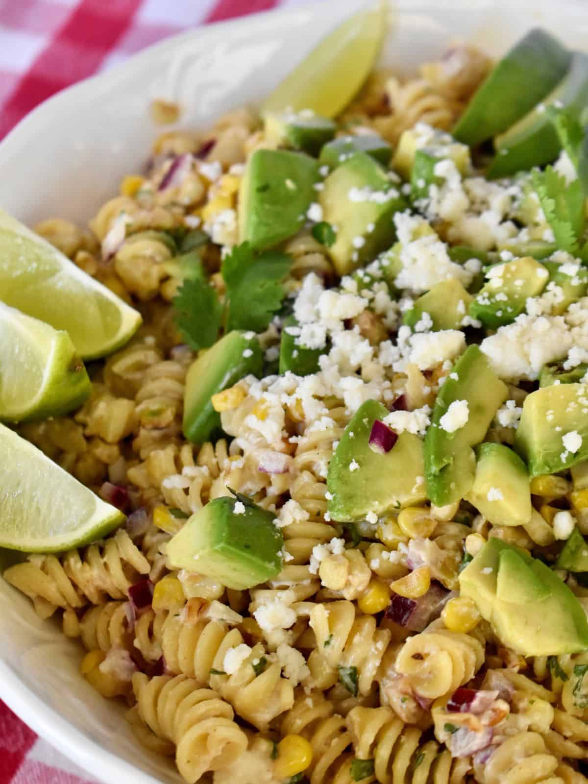 Elote Pasta Salad in a white bowl with lime wedges and avocado on top. 