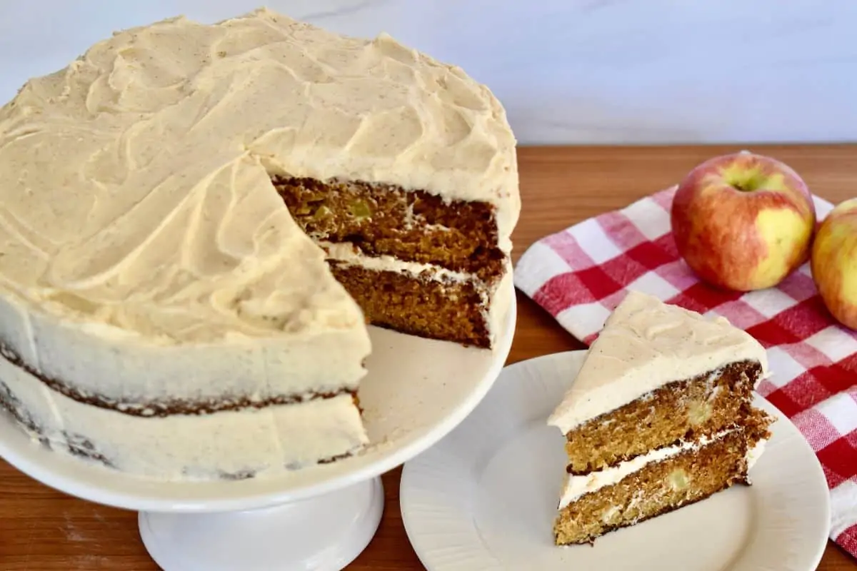 Apple spice cake with cinnamon buttercream frosting o a cake stand with apples in the background. 