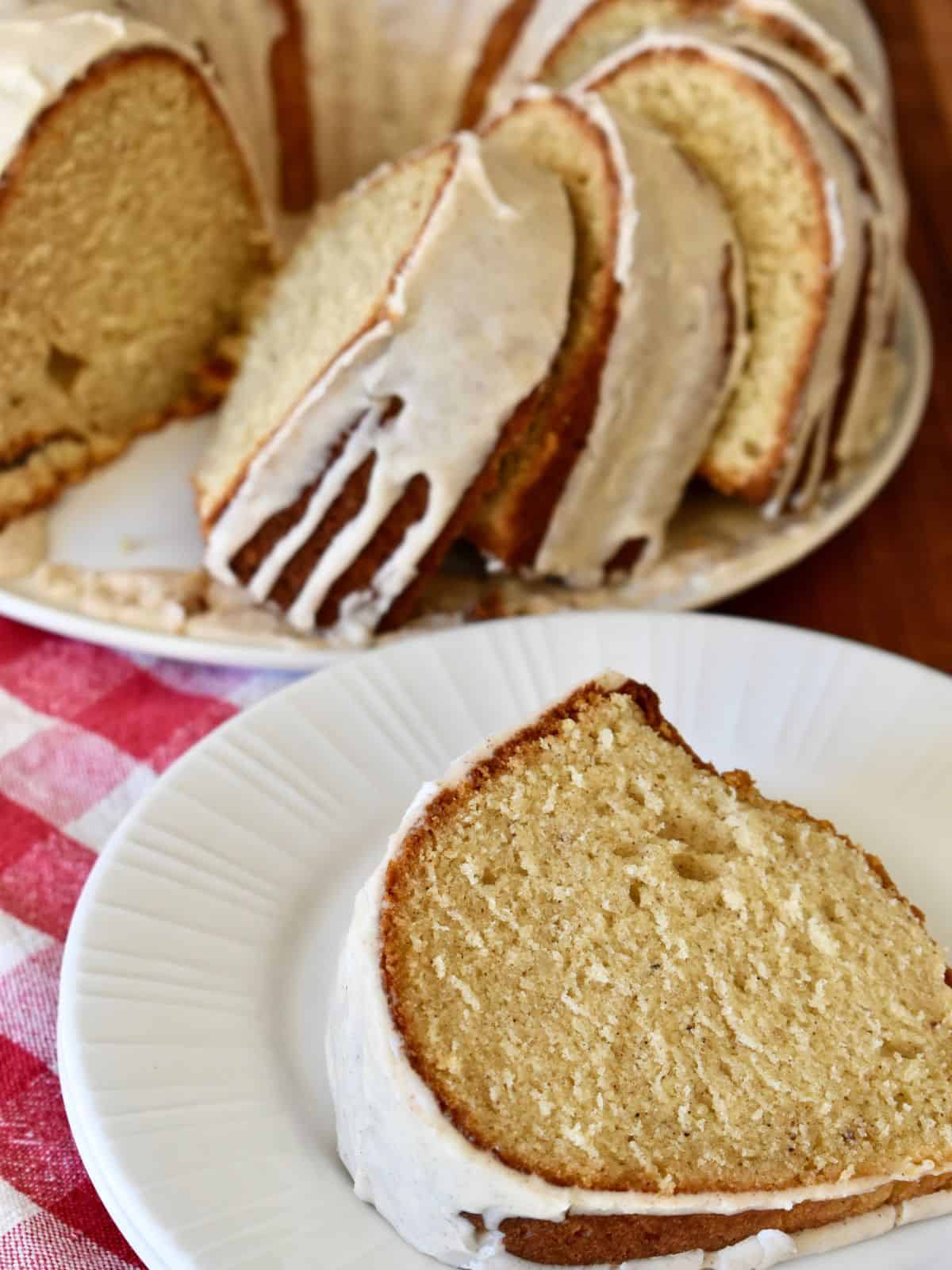 Slice of eggnog bundt cake on a white plate with the cake in the background. 