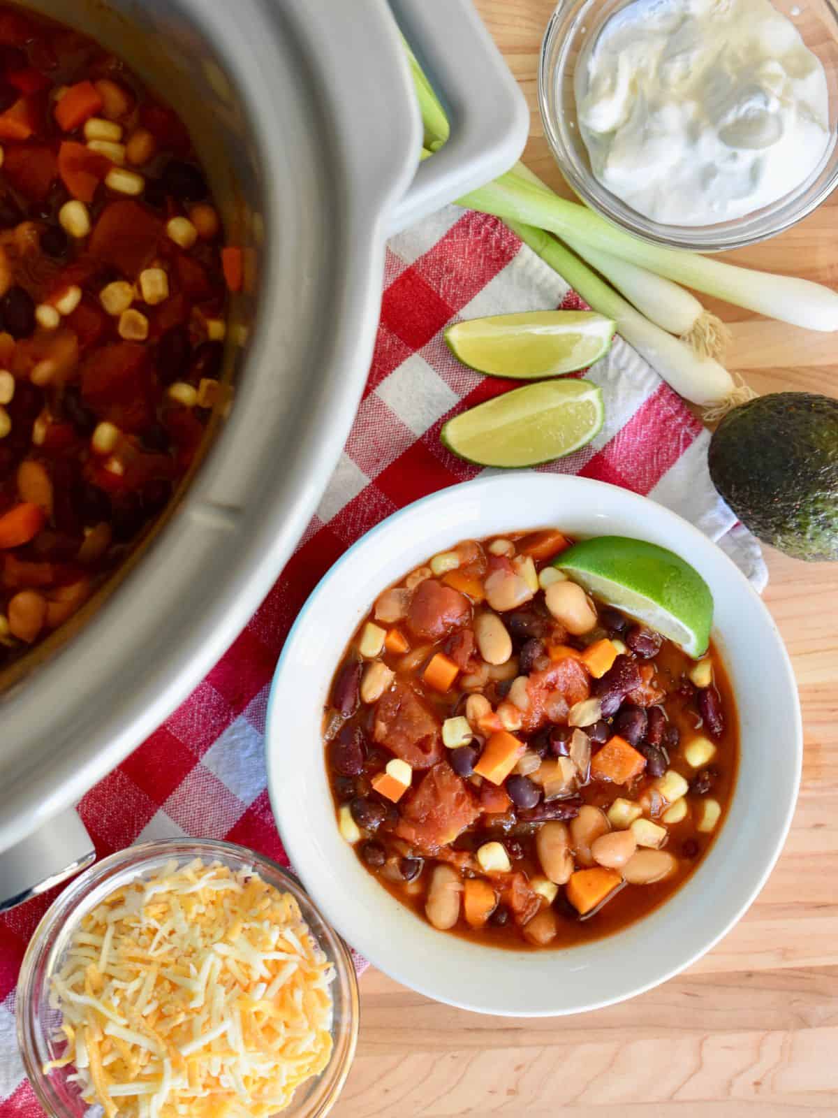 Three Bean Vegetarian Chili in a bowl with a lime wedge and slow cooker filled with chili next to it. 