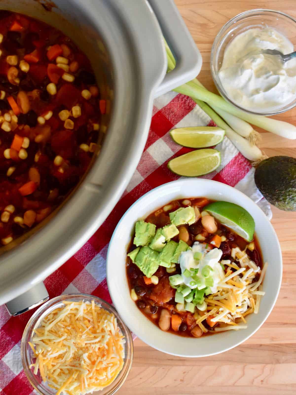 Three bean vegetarian chili in the slow cooker with sour cream, cheese, green onions, and avocado. 