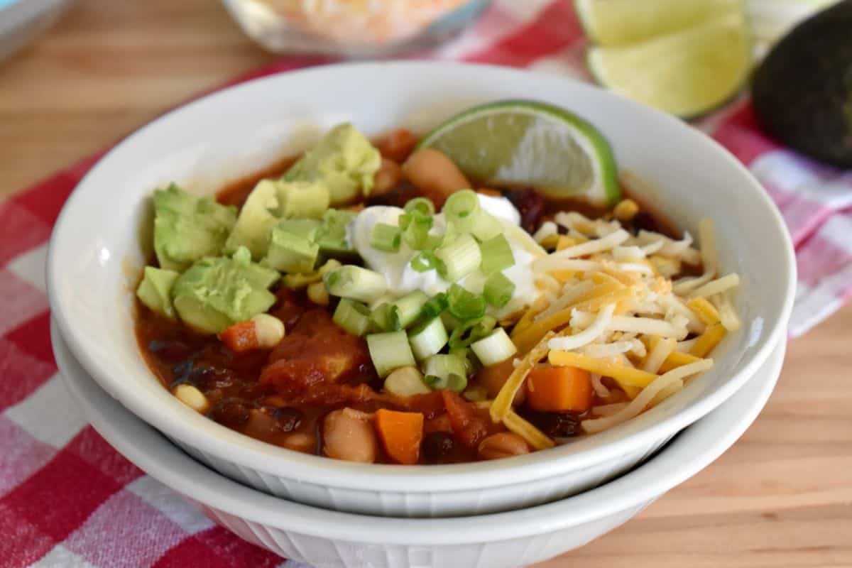 White bowl filled with three bean vegetarian chili topped with green onion, avocado, cheese, and a lime wedge. 