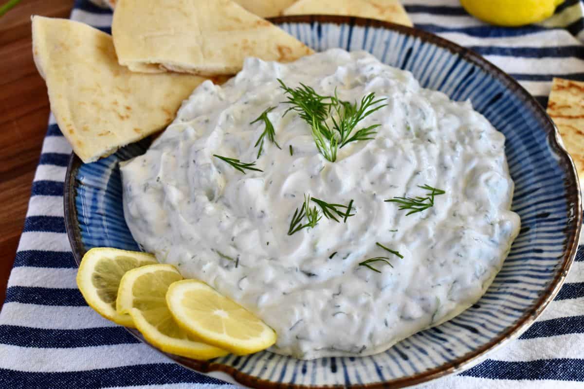Tzatziki sauce in a blue bowl with lemon wedges, dill, and pita bread. 