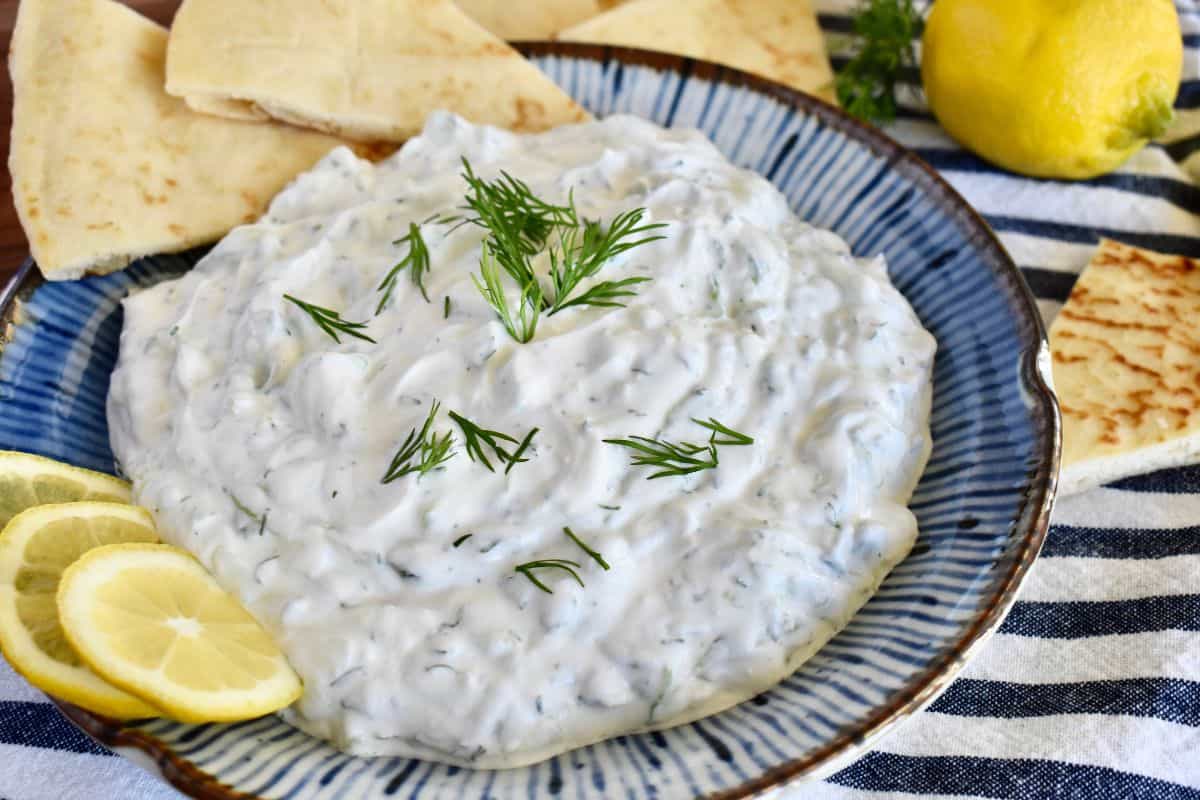 Tzatziki Sauce with Sour cream in a blue bowl with pita bread, lemon, and fresh dill. 