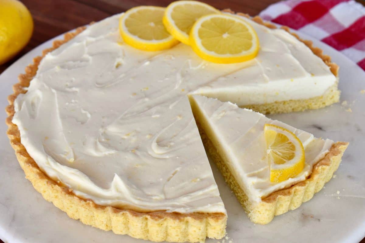 Slice of lemon mascarpone tart on a white plate with sliced lemon on top. 