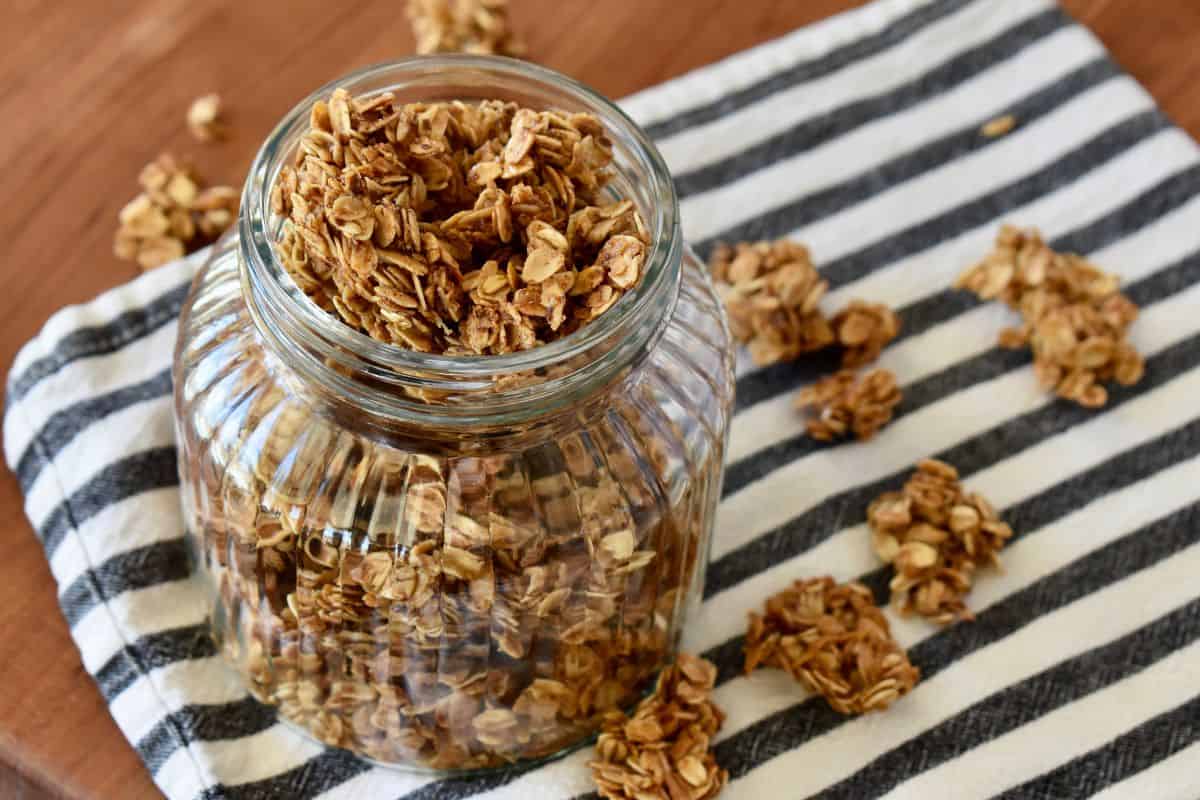 Granola in a glass container on a striped napkin with pieces of granola surrounding it. 