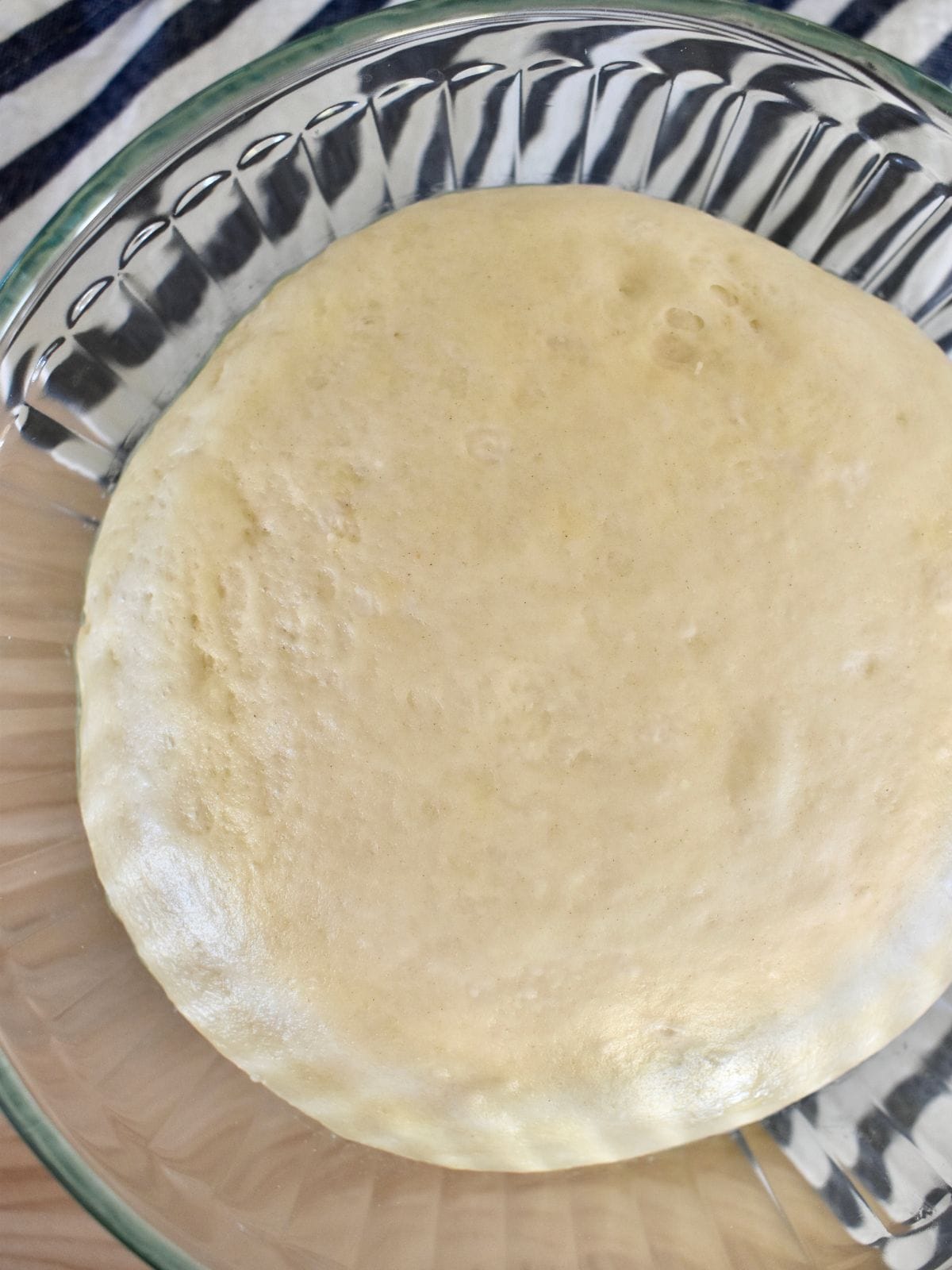 Pizza dough risen in a glass bowl on top of a striped napkin. 