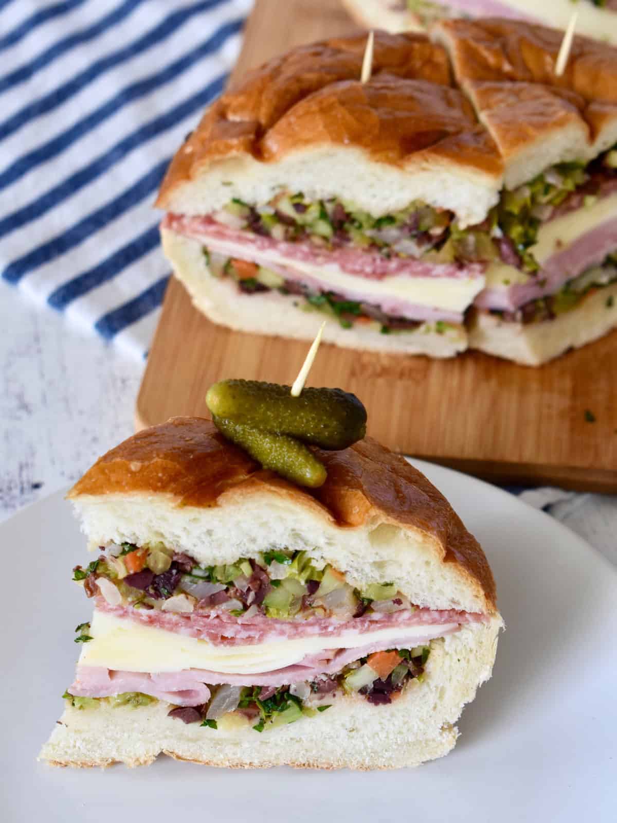 Slice of muffuletta sandwich on a white plate with pickles on top. 