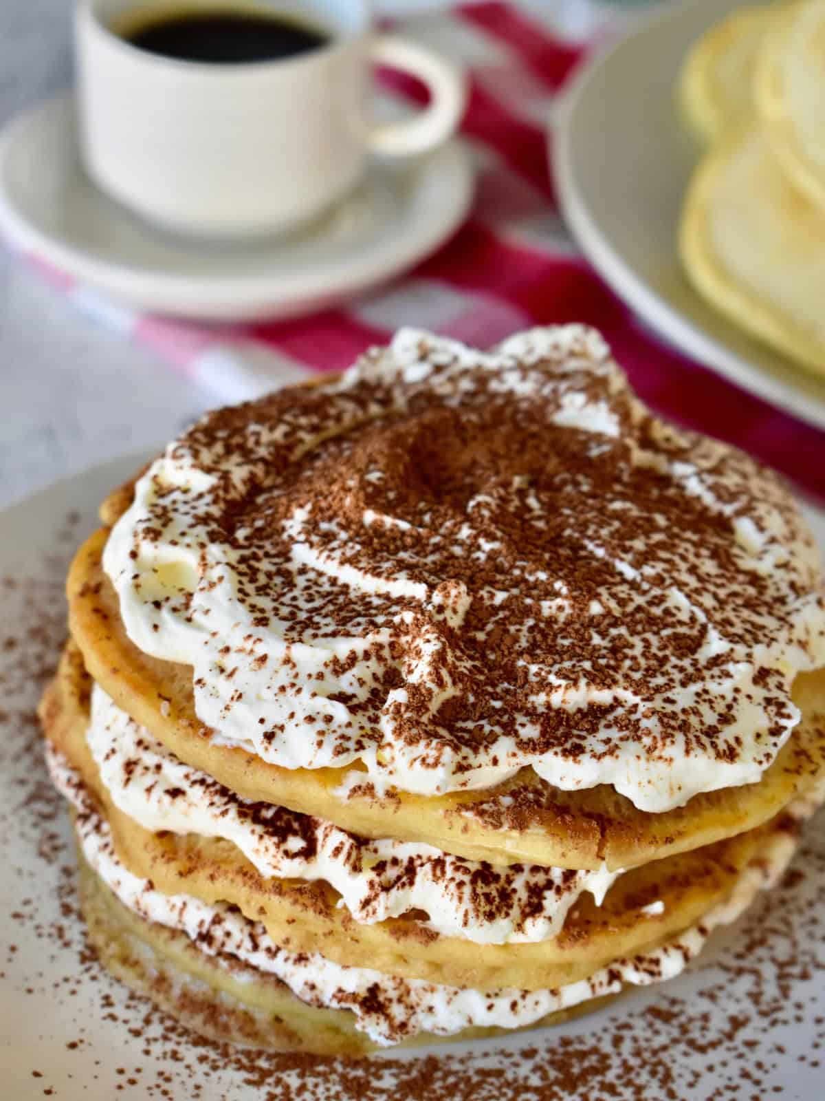 Tiramisu Pancakes on a white plate with a cup of coffee in the background. 