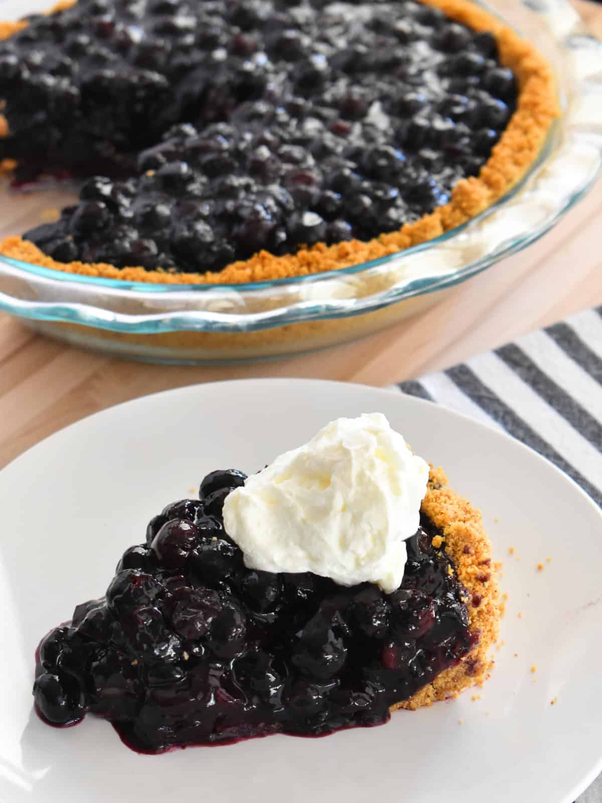 Blueberry pie with graham cracker crust on a white plate with whipped cream on top. 